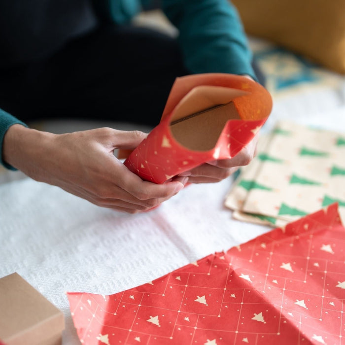 A person wraps a gift using red holiday paper decorated with small white trees. Other wrapping papers with green Christmas tree patterns, boxes, and ribbon are spread across the table, creating a festive gift-wrapping scene.