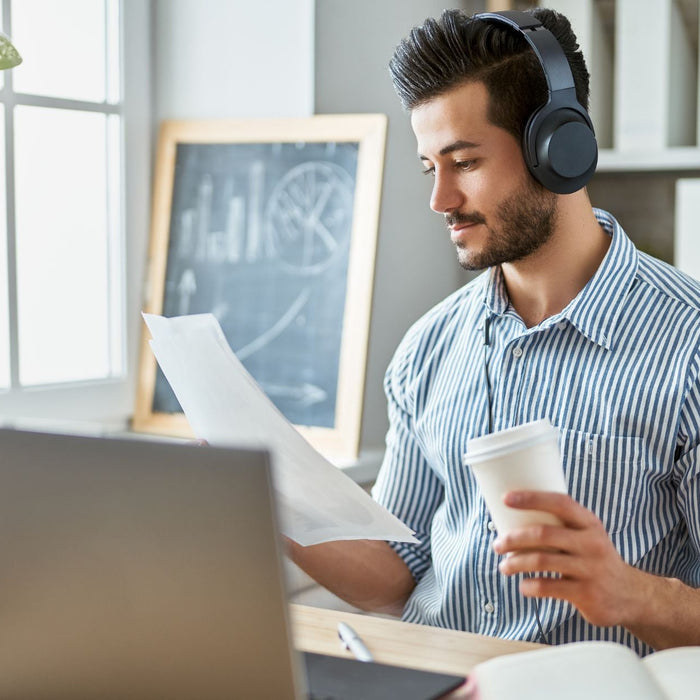 Man using workplace accommodations works remotely with headphones on, reviewing documents and holding a coffee cup at his desk, surrounded by a laptop, charts, and natural light in a plant-filled home office.