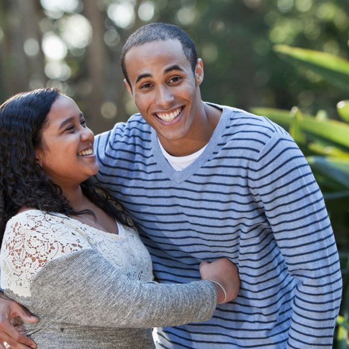 Smiling siblings stands close together outdoors, surrounded by lush green plants and trees. 