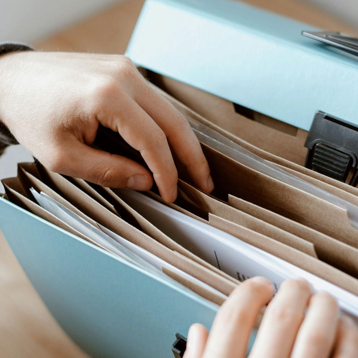 Close-up of hands organizing papers inside a blue accordion file folder. The folder’s brown dividers hold neatly arranged documents, suggesting organization or office work.