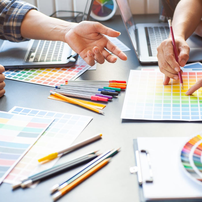Multiple people collaborate around a table reviewing printed color palettes, pointing at swatches while discussing design choices. Laptops, pens, and color guides are spread across the workspace, suggesting a branding or graphic design planning session.