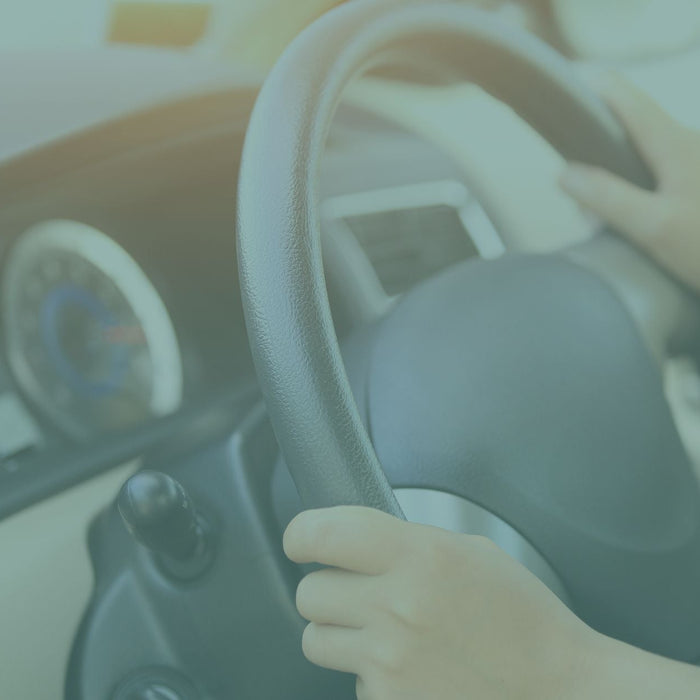 Close-up view of a person’s hands gripping a car steering wheel, with the dashboard and instrument cluster visible in the background. The softly lit image focuses on driving and vehicle control inside a car interior.