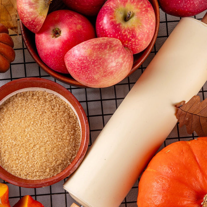 Overhead view of a fall arrangement featuring red apples, orange pumpkins, pumpkin slices, cinnamon sticks, brown sugar, mixed nuts, and a honey dipper. A cream colored rolling pin and dried autumn leaves complete the cozy seasonal baking scene.