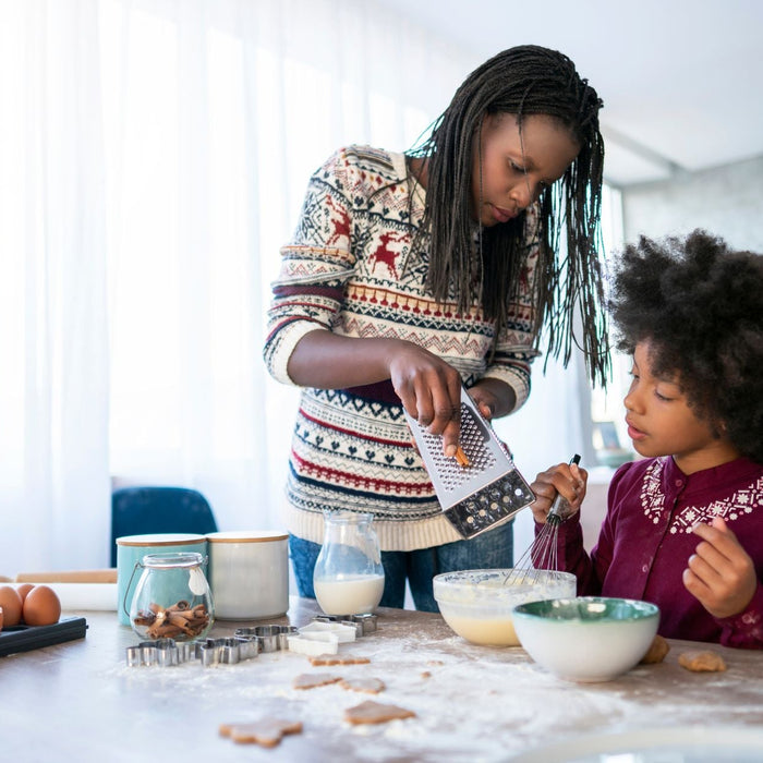 An adult and a child bake together in a bright kitchen. The adult is grating an ingredient into a mixing bowl.