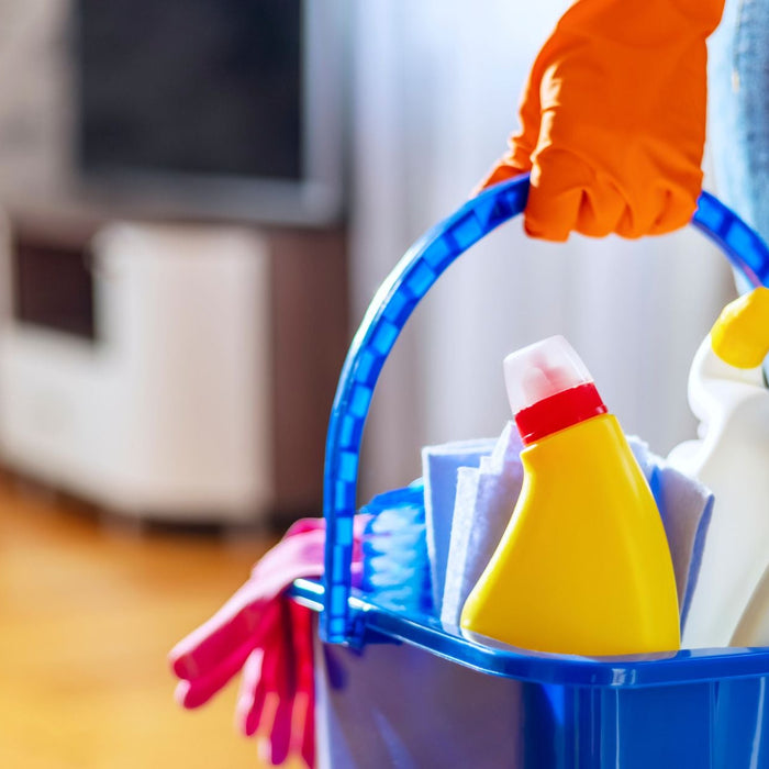 A person wearing orange gloves holds a blue plastic bucket filled with cleaning supplies, including spray bottles and cleaning cloths, in a brightly lit room.