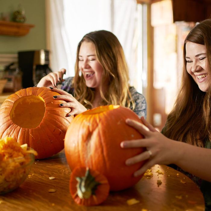 Two people laugh while carving pumpkins at a kitchen table, surrounded by pumpkin seeds and orange pulp. A bowl filled with pumpkin innards sits nearby, capturing a fun, messy autumn activity.