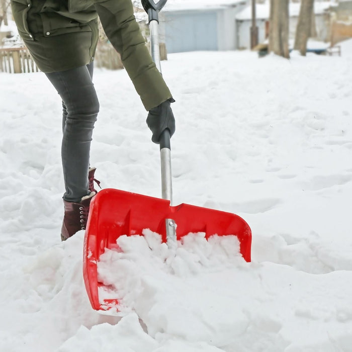 A person wearing winter boots and gloves uses a bright red snow shovel to clear a path through deep snow. Trees and houses are visible in the background, showing a typical winter day of outdoor cleanup.