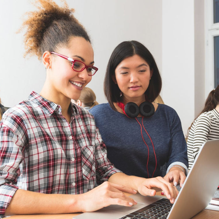 A group of young women are seated in a classroom working on laptops. Two of them, in the foreground, are collaborating and smiling while typing, with one wearing red glasses and a plaid shirt, and the other with headphones around her neck. Other students 