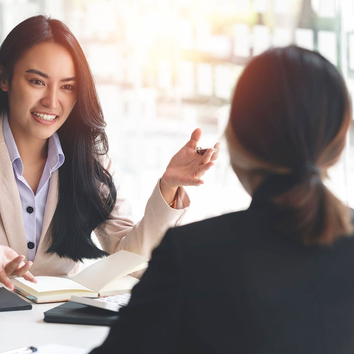 A woman in a light beige blazer smiles and gestures while speaking during a professional meeting or interview. She faces another person seated across the table, with notebooks and a calculator between them, and bright sunlight filters through large window