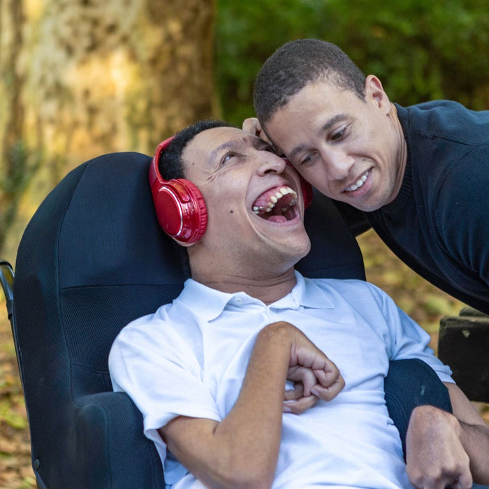 Two adults share a joyful moment outdoors in a leafy park. One person sits in a wheelchair wearing red headphones and laughs openly, while the other leans in close with a warm smile, suggesting a caring and affectionate interaction.