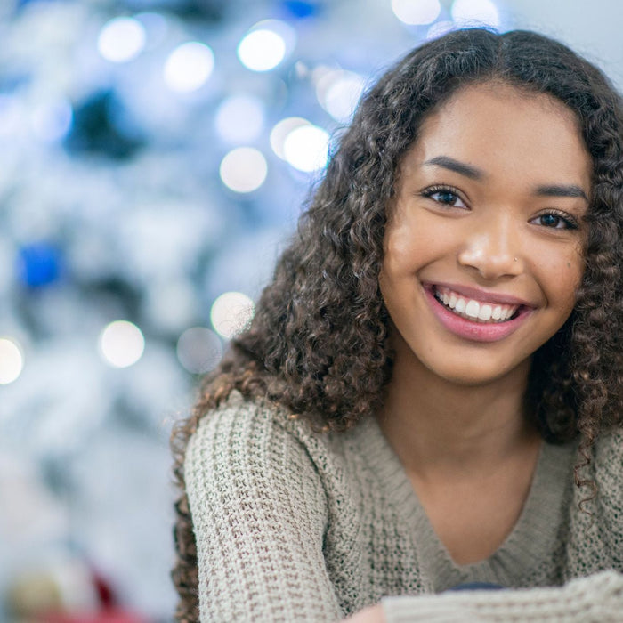 A young woman with long curly hair smiles warmly while sitting indoors, with soft holiday lights and a decorated Christmas tree blurred in the background.