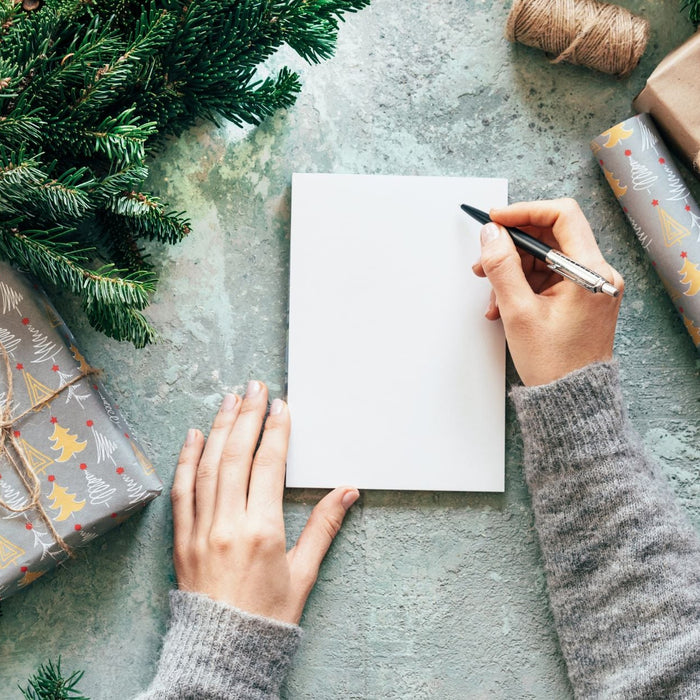 Hands write on a blank card laid on a textured surface, surrounded by wrapped holiday gifts, twine, and evergreen branches. The scene creates a cozy, festive flat-lay atmosphere, suggesting writing a Christmas or holiday greeting card.