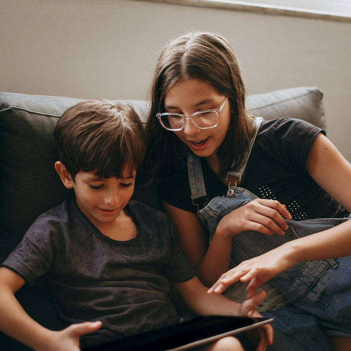 An older child and a younger child sit close together on a couch, looking at a tablet screen. The older child gestures as if explaining something, while the younger child watches attentively, creating a cozy, shared learning moment at home.