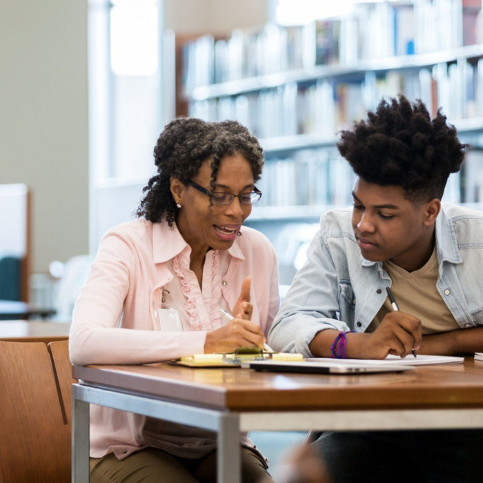 A teacher and student sit side by side at a table in a library, working through notes together. The teacher gestures while explaining, and the student listens closely with a pen in hand, surrounded by books and study materials.