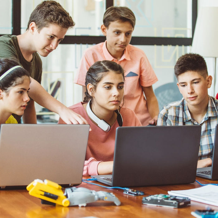 A group of teenagers collaborates around a table with open laptops, focusing intently on a project. Electronic parts and robotics equipment are spread across the table, suggesting a STEM or coding workshop.