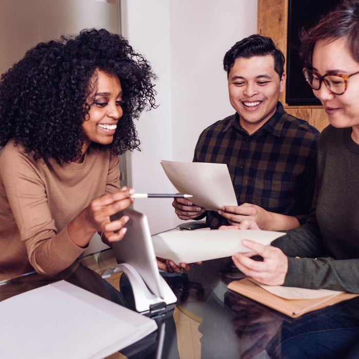 Three colleagues sit around a glass table in an office, smiling as they review documents together. One person holds a pen while pointing toward a tablet stand, creating a collaborative and positive work atmosphere.