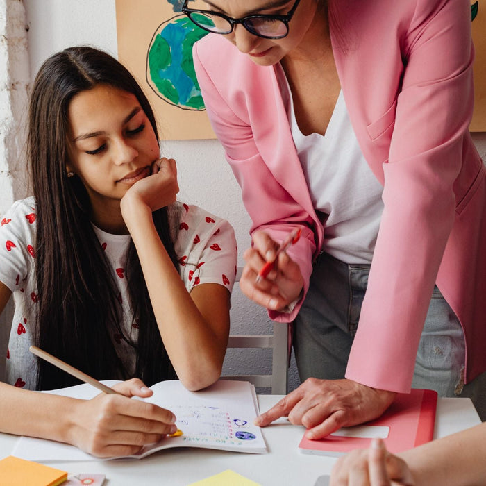 A student sits at a desk drawing in a notebook while a teacher in a pink blazer leans over, pointing and offering guidance. Another student works beside them, with colorful markers, sticky notes, and plants on the windowsill creating a bright, creative cl