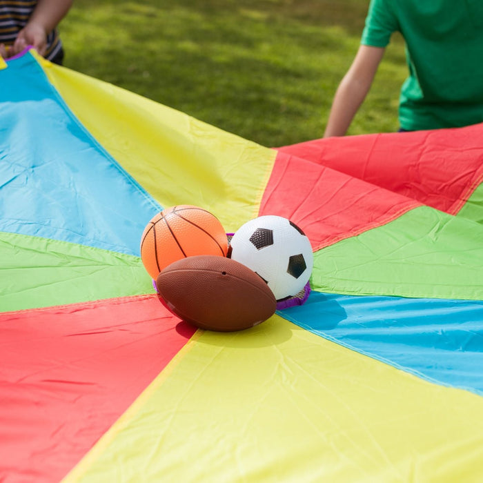 Children hold the edges of a colorful parachute game outdoors, with a basketball, soccer ball, and football balanced in the center. The bright activity encourages teamwork, coordination, and active play on a grassy field.
