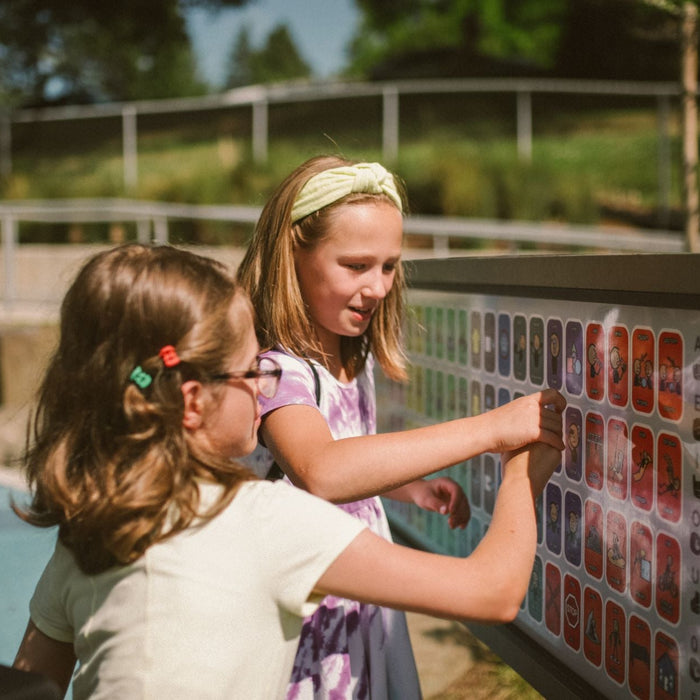 Two children interact with an outdoor educational activity board featuring letters, symbols, and colorful icons. 
