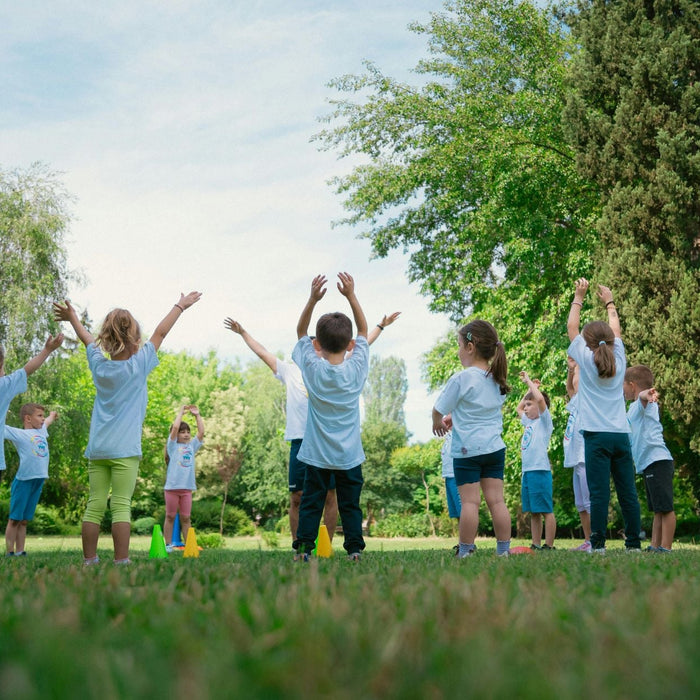 Group of young children stand in a circle on a grassy field with their arms raised during an outdoor activity led by an instructor. Surrounded by trees and colorful cones, the children participate in a physical education or team-building exercise in a par