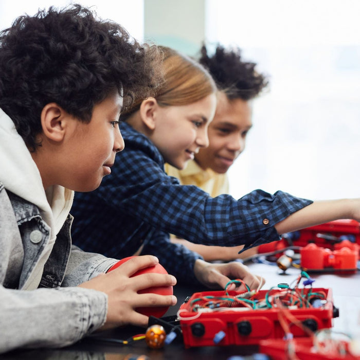 Three students collaborate on a robotics project, assembling and testing a small wheeled robot with exposed wiring and electronic components on a classroom table. Surrounded by robotics parts and tools, they work together with focused expressions during a