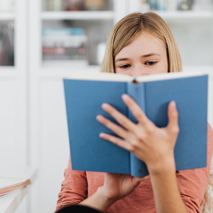 A young person holds a blue hardcover book up close to their face while reading indoors. A desk with notebooks and orange headphones sits nearby, and shelves with books and decor are visible in the softly lit background.