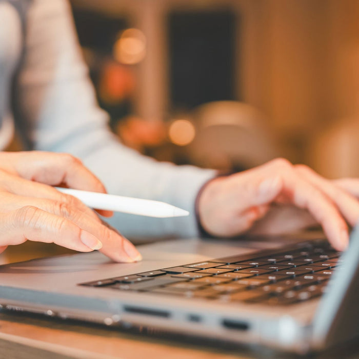 Close-up of hands using a laptop, with one hand typing on the keyboard and the other holding a stylus near the trackpad. Warm ambient lighting in the background creates a cozy workspace atmosphere, suggesting multitasking or digital design work.