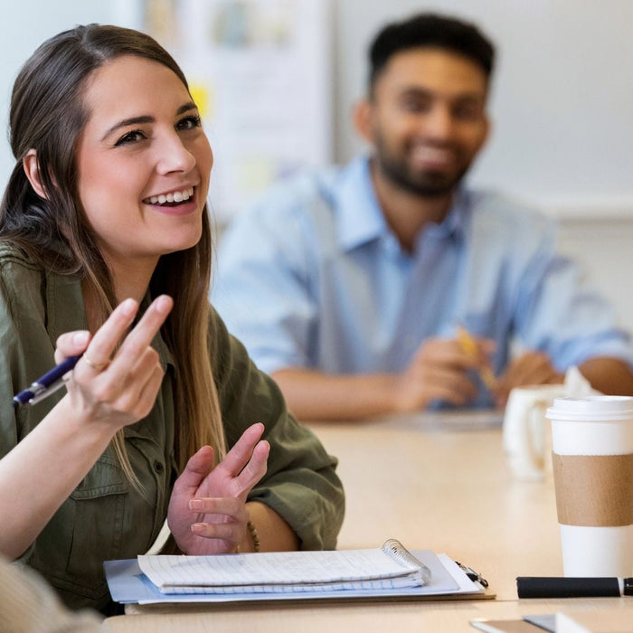 A smiling woman gestures with a pen while participating in a group discussion around a conference table. Several colleagues listen in the background, with notebooks, a laptop, and a coffee cup on the table during the collaborative meeting.