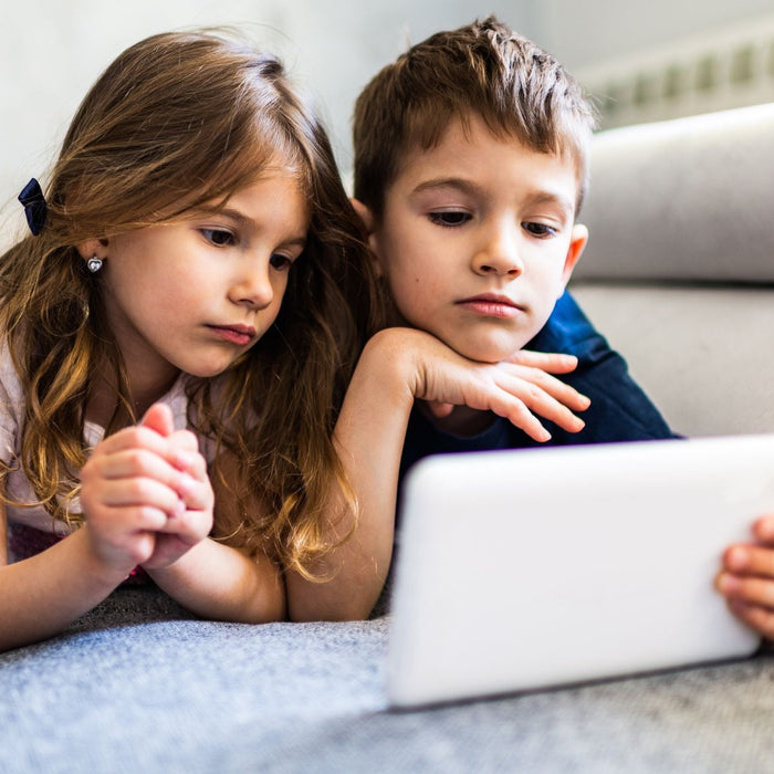 Two young children lie side by side on a couch, focused on a tablet screen as they watch or play together. The girl and boy rest their heads on their hands, showing concentration in a cozy indoor setting.
