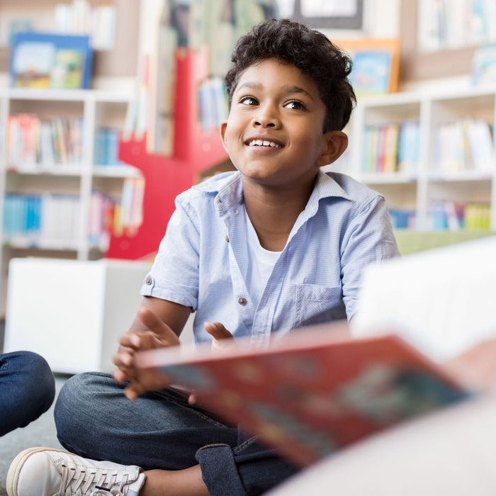 Group of children sit together in a library or classroom during a group discussion, with one smiling boy speaking while classmates listen nearby. Bookshelves filled with colorful books create a bright and collaborative learning environment.