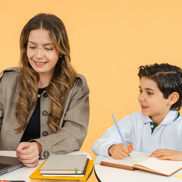 Teacher or tutor sits beside a young student, using a laptop while the child writes in a notebook during a lesson or tutoring session. The pair work together at a desk with school supplies and notebooks against a simple yellow background.