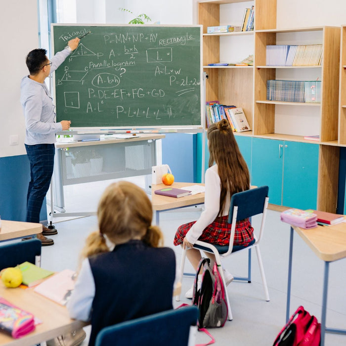 Teacher writes geometry formulas and diagrams on a chalkboard while students sit at desks facing forward in a bright classroom. Bookshelves line the wall, and students follow along with notebooks during a math lesson.