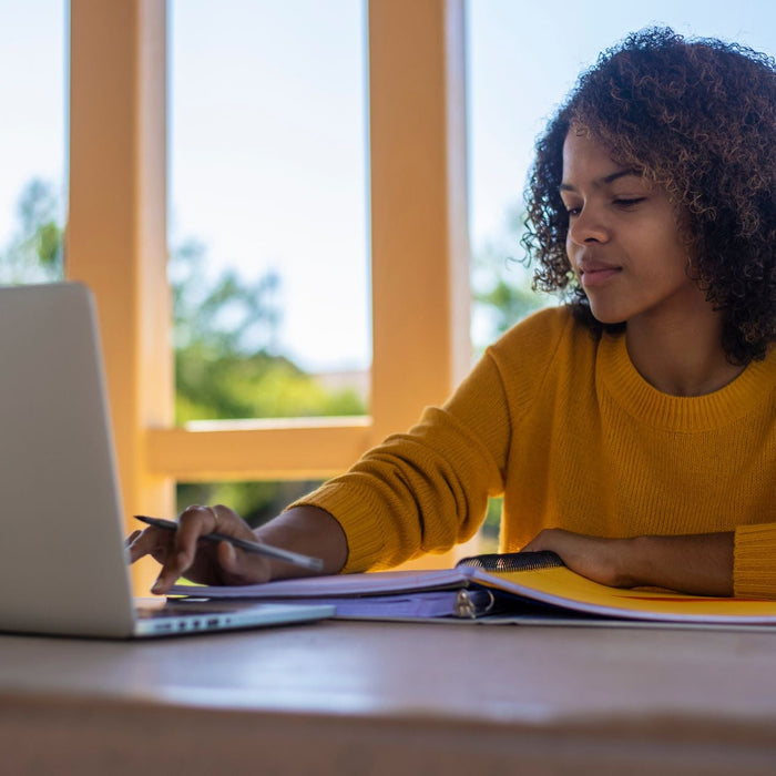 A young woman in a yellow sweater studies outdoors at a table, working between a laptop and an open binder. Sunlight and greenery surround the space, creating a bright and focused study environment.