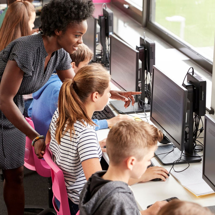 A teacher leans over to assist a student at a row of desktop computers in a classroom. Several students sit side by side working on assignments, while the teacher points at a screen, offering guidance and support.
