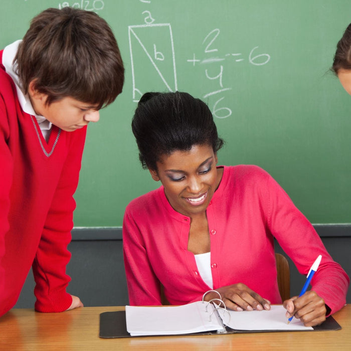 A smiling teacher sits at a desk in front of a chalkboard, reviewing notes with two students who are leaning in attentively. 