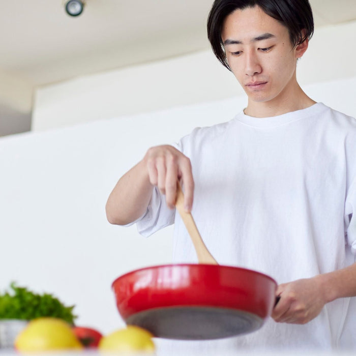 A young adult stands in a bright kitchen stirring food in a red frying pan with a wooden spoon. Fresh ingredients like lemons and leafy greens sit on the counter nearby, suggesting a healthy home-cooked meal in progress.