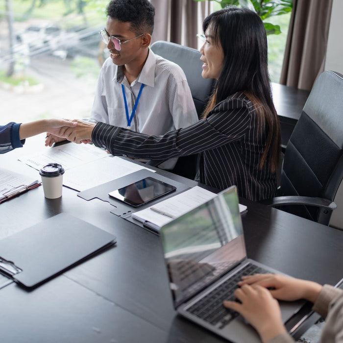 A job interview takes place in a modern office as a woman in a navy blouse shakes hands with an interviewer across the table. Three interviewers sit with documents, laptops, and coffee cups, smiling as they discuss and review the candidate’s resume.